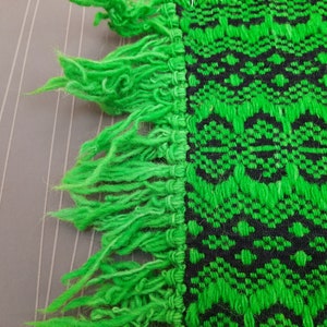 May include: Green and black woven fabric with a fringe border. The fabric has a geometric pattern.