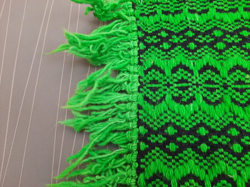 May include: Green and black woven fabric with a fringe border. The fabric has a geometric pattern.