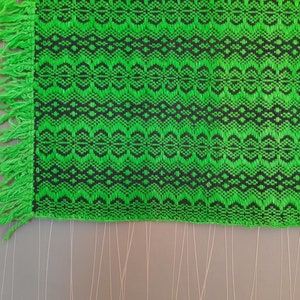 May include: Green and black woven fabric with a fringe border. The fabric has a geometric pattern.