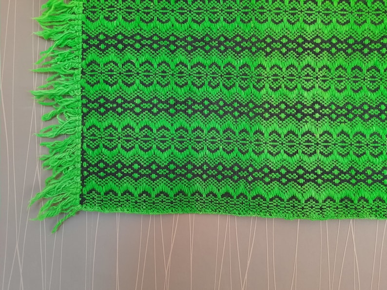 May include: Green and black woven fabric with a fringe border. The fabric has a geometric pattern.