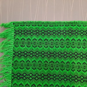 May include: Green and black woven fabric with a fringe border. The fabric has a geometric pattern.