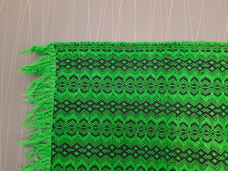 May include: Green and black woven fabric with a fringe border. The fabric has a geometric pattern.