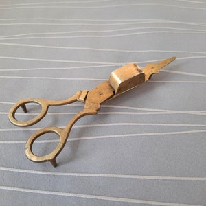 May include: Antique brass candle snuffer with a scissor-like design. The snuffer has a small, rectangular compartment on the top for holding a candle wick.