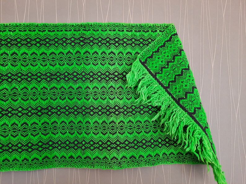May include: A green and black woven rug with fringe. The rug has a geometric pattern.