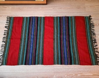 Puede incluir: Una alfombra de rayas rojas, verdes, azules y negras con flecos en los bordes.