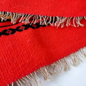 Pu&ograve; includere: Tappeto rosso tessuto con un motivo geometrico bianco e nero e un bordo a frangia.