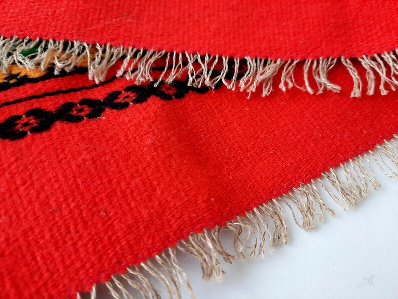 Pu&ograve; includere: Tappeto rosso tessuto con un motivo geometrico bianco e nero e un bordo a frangia.