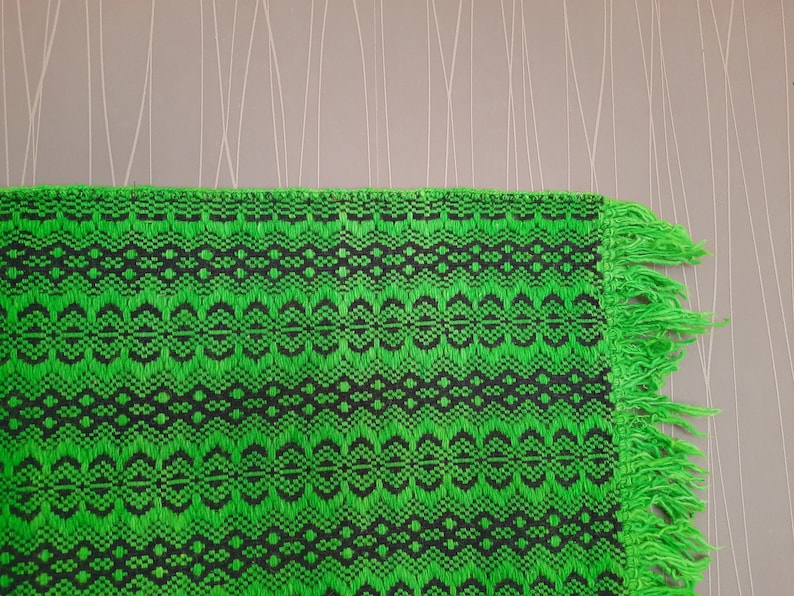 May include: Green and black woven rug with fringe on the edges. The rug has a geometric pattern.