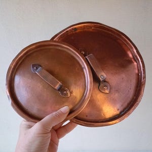 Puede incluir: Dos tapas de olla de cobre con asas. La tapa más pequeña está en primer plano y la más grande está detrás.