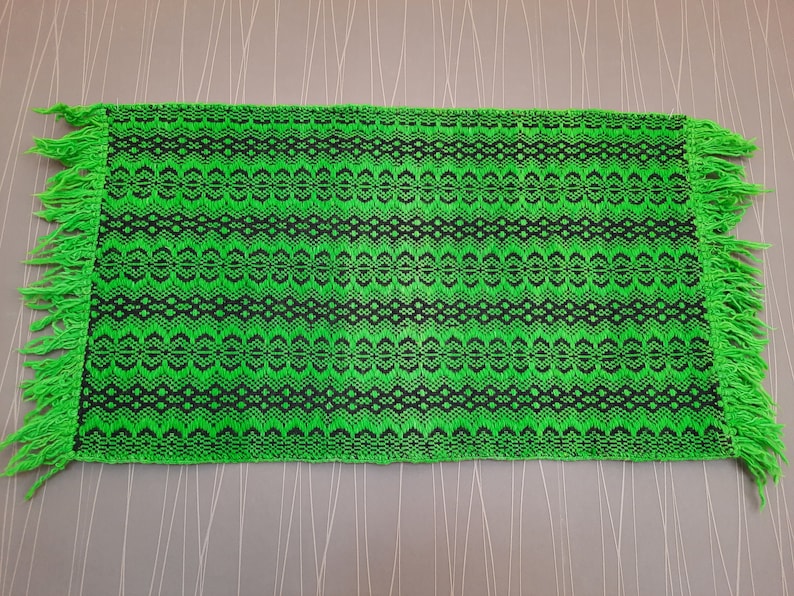 May include: A green and black woven rug with fringe on all sides. The rug has a geometric pattern.