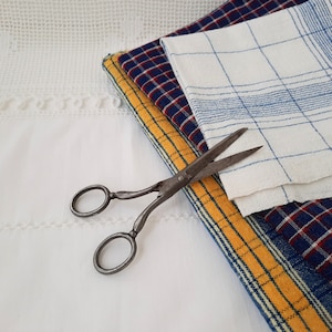 May include: A pair of antique scissors with a yellow, blue and red plaid fabric underneath. The fabric is folded in thirds.