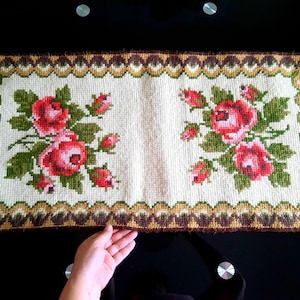 Puede incluir: Una alfombra rectangular de color crema con un borde marrón y dorado. La alfombra está bordada con rosas rojas y verdes.