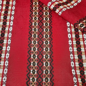 Puede incluir: Textil rojo con intrincados patrones tejidos. El diseño presenta rayas verticales con motivos geométricos y florales en blanco, negro y marrón. La tela está doblada, mostrando la detallada artesanía.