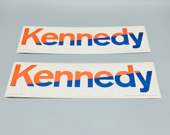 May include: Two white rectangular stickers with the word "Kennedy" printed in orange and blue letters. The word is split in half with the top half in orange and the bottom half in blue.