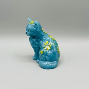 May include: A ceramic cat figurine in a seated position. The cat is a vibrant teal color with yellow and white floral accents. The figurine has a glossy finish and is a decorative item.
