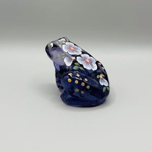 May include: A ceramic frog figurine in a sitting position. The frog is dark blue with white and pink floral designs on its back and gold dots on its sides. The figurine has a glossy finish.
