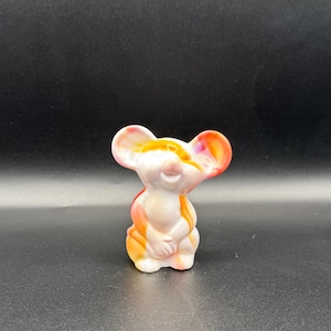 May include: A small, ceramic mouse figurine with a smiling face and folded paws. The mouse has large, rounded ears and is painted with a blend of white, orange, and pink hues. The figurine is standing upright against a dark background.