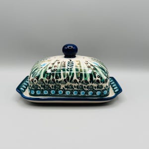 May include: A rectangular ceramic butter dish with a lid featuring a dark blue knob. The surface is decorated with a hand-painted pattern of green leaves, small brown dots, and light blue circular accents on a cream-colored background.