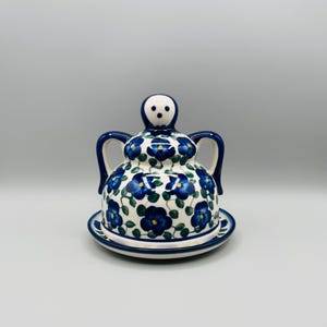 May include: A ceramic butter dish with a lid and a matching plate. The dish is white with a blue floral pattern and dark blue accents. The lid has a handle and two side handles.