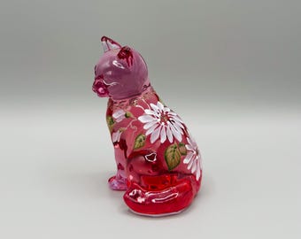 May include: A decorative glass cat figurine in shades of pink and red. The cat is sitting with its tail curled and features white daisy floral designs on its body. The figurine is a unique home decor item.