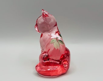 May include: A pink glass cat figurine with a floral design. The cat is sitting, with its head turned to the side. The floral design features white flowers and green leaves. The glass has a glossy finish.