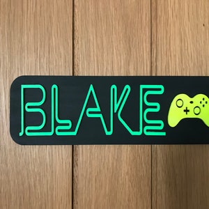 May include: A black rectangular sign with rounded corners features the name "BLAKE" in neon green, stylized lettering. A neon green video game controller is on the right. The sign is mounted on a wooden background.