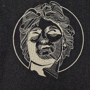 May include: Embroidered design of a woman's face with her eyes closed, surrounded by a circle, on a dark blue fabric.