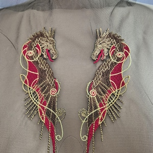Puede incluir: Dos apliques bordados de dragones. Cada dragón presenta una cabeza detallada, cuerpo marrón y dorado, y detalles rojos en forma de alas. Los apliques están colocados sobre un fondo de tela gris, creando un diseño simétrico.
