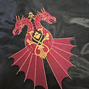 Op de afbeelding: Geborduurde patch met een tweekoppige rode draak met gouden accenten. De draak staat op een cirkelvormig ontwerp met vleugelachtige elementen, tegen een zwarte achtergrond. De borduursels zijn gedetailleerd en ingewikkeld.