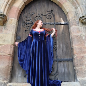 Könnte beinhalten: Eine Frau in einem blauen Samtkleid mit langen roten Haaren steht vor einem gewölbten Tor. Das Kleid hat lange Ärmel und einen roten Saum am Ausschnitt. Das Tor ist aus Stein und hat eine Holztür mit Eisenverzierungen.