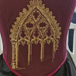 Puede incluir: Corsé burdeos con diseño de arco gótico bordado en oro. El corsé presenta un corpiño estructurado con costuras verticales y una falda negra en contraste. Un fino ribete magenta perfila la cintura.