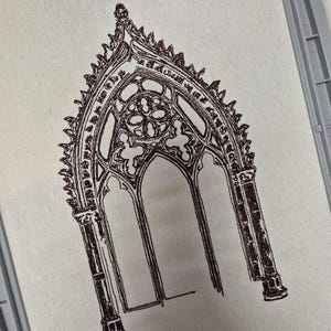 Puede incluir: Dibujo lineal marrón de una ventana gótica arqueada sobre tela color crema. La ventana presenta tracería detallada y tres aberturas arqueadas. El diseño recuerda la arquitectura de las catedrales.