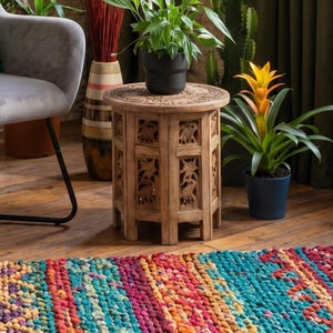 May include: A living room interior featuring a grey armchair with a green cushion, a small carved wooden table, and various potted plants. The room has a colourful rug and olive green curtains.
