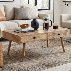May include: A living room scene with a beige sofa, cushions, a wooden coffee table, and a woven leather ottoman. The room includes a rug, artwork, and a black floor lamp, creating a stylish interior. The coffee table is approximately 120 cm long.