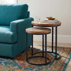 May include: Two round nesting side tables with wooden tops and black metal frames. The larger table has a small gold bowl and books on top. The tables are on a patterned rug, next to a teal sofa.