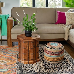 May include: A wooden octagonal side table with metal accents sits on a colourful rug in a living room. A large green plant in a black pot sits on the table. A colourful patterned pouffe sits on the rug in front of the table.