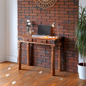 May include: A wooden console table with three drawers and turned legs. The table is made of dark wood and has a distressed finish. It is sitting in front of a brick wall with a lamp on the table.