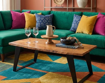 May include: A living room scene featuring a green velvet sectional sofa with colorful throw pillows in yellow, fuchsia, and navy blue. A wooden coffee table with black metal legs sits in front of the sofa, with wine glasses, a pitcher, and a decorative bowl.