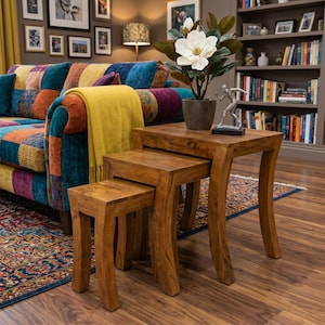 May include: A set of three wooden nesting tables in a warm brown hue. The tables have curved legs and a square top. The largest table supports a potted plant with a large white flower. The tables are in a living room setting, with a sofa in the background.