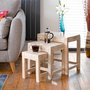 May include: Three white wooden nesting tables with a coffee cup and a French press on the top table. The tables are in front of a grey sofa with colourful cushions.