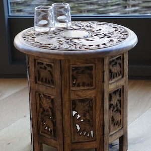 Puede incluir: Una peque&ntilde;a mesa auxiliar de madera tallada con una parte superior redonda y un cuerpo cil&iacute;ndrico. La mesa presenta tallas en relieve detalladas de elefantes y motivos florales. Dos vasos transparentes est&aacute;n encima. La mesa mide aproximadamente 61 cm de alto.