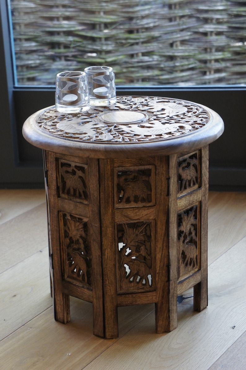 Puede incluir: Una peque&ntilde;a mesa auxiliar de madera tallada con una parte superior redonda y un cuerpo cil&iacute;ndrico. La mesa presenta tallas en relieve detalladas de elefantes y motivos florales. Dos vasos transparentes est&aacute;n encima. La mesa mide aproximadamente 61 cm de alto.
