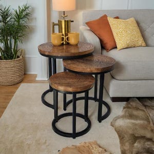 May include: A trio of nesting tables. Each table has a round, carved wooden top and black metal legs. The tables are of varying heights and sizes, with the largest featuring a detailed circular design. The tables are on a light-coloured rug.