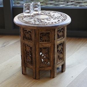 Puede incluir: Una peque&ntilde;a mesa auxiliar redonda de madera con intrincados detalles tallados. La superficie de la mesa presenta un dise&ntilde;o circular con motivos florales y animales. Los lados de la mesa est&aacute;n decorados con paneles tallados. Dos vasos transparentes est&aacute;n sobre la mesa.