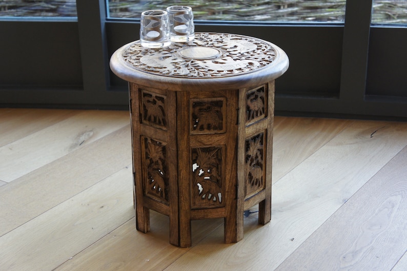 Puede incluir: Una peque&ntilde;a mesa auxiliar redonda de madera con intrincados detalles tallados. La superficie de la mesa presenta un dise&ntilde;o circular con motivos florales y animales. Los lados de la mesa est&aacute;n decorados con paneles tallados. Dos vasos transparentes est&aacute;n sobre la mesa.