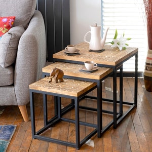 May include: A set of three nesting tables with a wood top and black metal frame. The tables are decorated with an intricate carved design. The top table has a small elephant figurine on it.