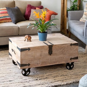 May include: A light wood coffee table with black metal accents and wheels. The table has a rectangular top and a storage compartment. A small wooden elephant figurine and a potted plant with yellow flowers are on the table.