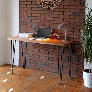 May include: A wooden desk with black hairpin legs. A laptop, books, a small lamp, and a mug of tea are on the desk. The desk is in front of a red brick wall. A plant in a white pot is to the right.