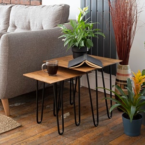 May include: Two wooden nesting tables with black hairpin legs. A cup of coffee and an open book rest on the tables. The tables are in front of a grey sofa. Plants and decorative items are in the background.