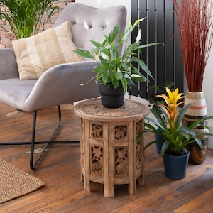 Puede incluir: Un sal&oacute;n acogedor con un sill&oacute;n gris, una mesa auxiliar de madera tallada y varias plantas en macetas. La mesa tiene intrincadas tallas de elefantes. Una alfombra tejida y suelos de madera completan el espacio.